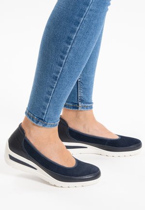 Marineblaue Slipper mit Wildlederoberfläche, einer weißen Keilsohle und abgerundeter Spitze. Beine in engen blauen Jeans sichtbar.