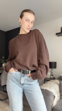 Barna, oversized pulóver, ejtett vállakkal, könnyű kötött anyagból, világos mosású farmernadrággal párosítva és egy fekete övvel, ezüst csattal.