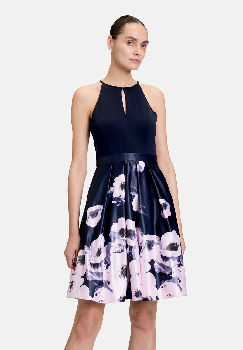 Femme portant une robe bleu marine sans manches avec un décolleté en forme de trou de serrure et une jupe à motif floral dans des tons de rose et noir.