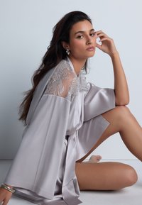 Robe de nuit en satin gris clair avec des détails en dentelle sur les épaules, dotée de manches amples et d'une ceinture à nouer, présentée sur un fond uni.