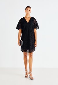 Μαύρο δαντελένιο φόρεμα με v-neck και φουσκωτά μανίκια, που διαθέτει περίτεχνα σχέδια και μήκος λίγο πάνω από το γόνατο. Συνδυάζεται με σανδάλια με λουράκια.