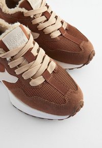 Scarpe da ginnastica marroni con tomaia in rete e camoscio, lacci beige e suola bianca. Presentano linguetta con marchio e dettagli testurizzati.