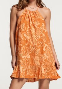 Woman wearing sleeveless orange halter dress with beige floral patterns and ruffled hem, standing against a plain light background.
