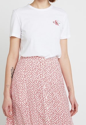 Osoba nosząca białą koszulkę Calvin Klein włożoną w białą spódnicę z małym czerwonym wzorem kwiatowym, pokazująca tatuaże na obu przedramionach.