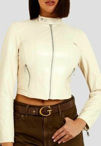 Woman wearing a fitted cream leather jacket with zipped pockets and a brown belt over dark pants, hand resting on hip.