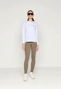 Λευκό πουκάμισο με μακριά μανίκια και στρογγυλή λαιμόκοψη, με μικρό λογότυπο, συνδυασμένο με καφέ κολάν. Το μοντέλο φοράει μαύρα γυαλιά ηλίου και μπεζ αθλητικά παπούτσια.