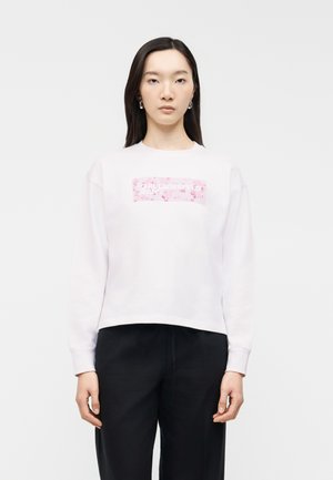 Młoda kobieta z długimi ciemnymi włosami ubrana w białą bluzę z różowym kwiatowym prostokątnym logo i czarnymi spodniami, stojąca przodem na białym tle.