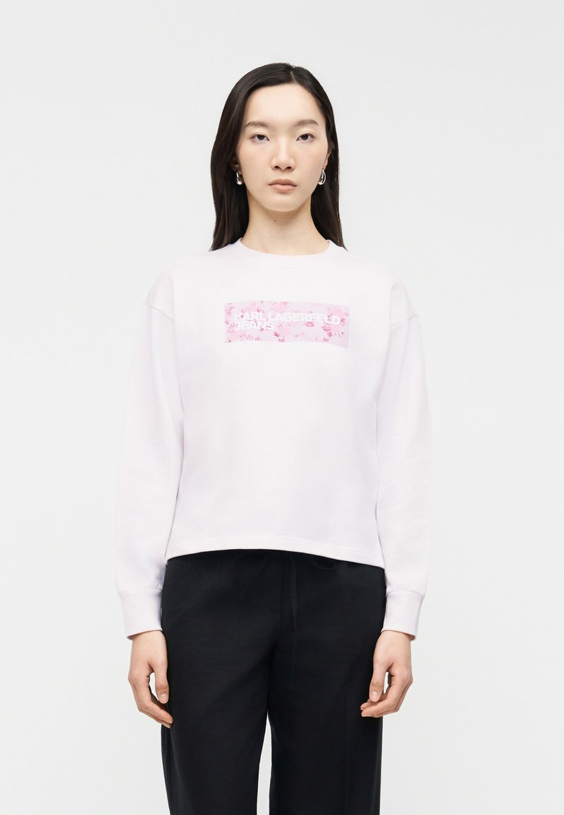 Νεαρή γυναίκα με μακριά σκούρα μαλλιά, ντυμένη με λευκή μπλούζα φούτερ που έχει ροζ ορθογώνιο λογότυπο με λουλούδια και μαύρο παντελόνι, κοιτάζοντας μπροστά σε λευκό φόντο.