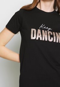 Camiseta negra de algodón con la frase "Sigue BAILANDO" en purpurina metálica color rosa dorado. Mangas cortas y un cuello redondo clásico.