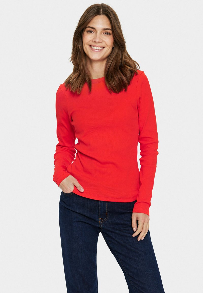 Sarkans longsleeve tops ar apaļu dekoltē un ribotu tekstūru, kombinēts ar tumši ziliem džinsiem. Modelis pozē ar vienu roku kabatā.