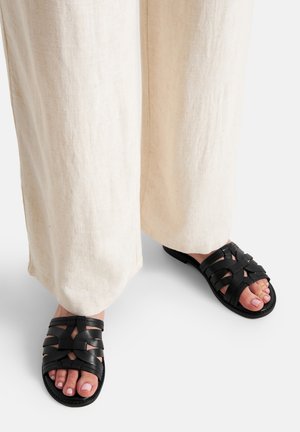 Czarne sandały ze skóry z plecionymi paskami i odkrytymi palcami, zestawione z beżowymi, luźnymi spodniami z lnu.