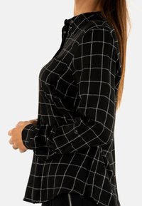 Camisa negra de botones con un patrón de cuadrícula blanca, mangas largas con puños abotonados, y una tela suave con un corte relajado.
