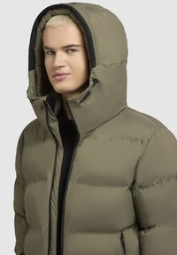 Pūdera jaka, olīvzaļā krāsā, ar kvadrāta tekstūru, augsts apkaklis ar regulējamiem sasienamiem aukliņām, melni akcenti, priekšējā rāvējslēdzēja aizdare, elegants dizains.