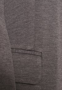 Harmaa kangas, jossa on teksturoitu kudonta ja taskudetalji. Materiaali on sileä ja viimeistelty.