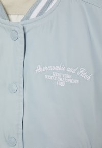 Lichtblauwe jas met een gladde textuur, voorzien van een geribde kraag met witte strepen en een geborduurd logo: "Abercrombie and Fitch" en "1892."