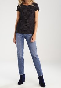 Kobieta ubrana w czarną koszulę z krótkim rękawem w kropki, jasnoniebieskie dżinsy oraz ciemnoniebieskie botki na wysokim obcasie, stojąca na jednolitym tle.