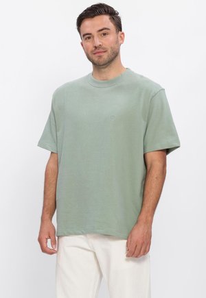 Man met kort donker haar, die een lichtgroen oversized T-shirt met korte mouwen en witte broek draagt, staand tegen een effen witte achtergrond.