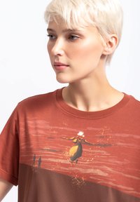Roestkleurig katoenen t-shirt met een afbeelding van een vliegende figuur tegen een zonsondergang achtergrond, met een soepele textuur en een ronde halslijn.