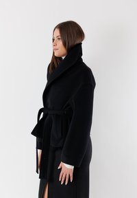 Cappotto avvolgente nero realizzato in tessuto spesso, con un ampio colletto, cintura in vita e maniche lunghe. Texture liscia con un design minimalista.