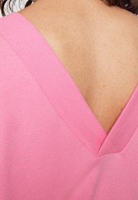 Różowy top z dekoltem w serek, wyróżniający się prążkowaną teksturą i głębokim, skośnym dekoltem. Tkanina wydaje się lekka i gładka, podkreślając prostotę.