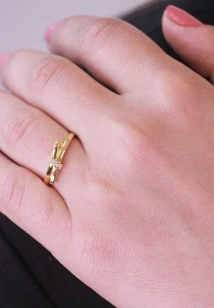 Gouden ring met een kruisontwerp,accenten van sprankelende stenen, gedragen op een hand met een lichte huidtint en een subtiele textuur.