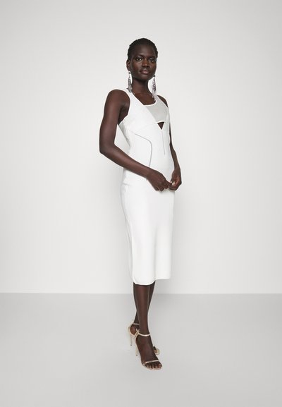 Donna in piedi con un vestito midi bianco senza maniche e dettagli cut-out, che indossa orecchini pendenti e sandali con tacco alto dorati.