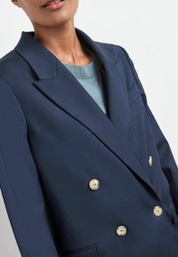 Námořnické modré dvouřadé sako se zlatými knoflíky, ostrými klopami a texturovanou tkaninou. Obsahuje detail náprsní kapsy.