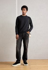 Young man wearing black sweatshirt, dark jeans, black sneakers with white soles, carrying small beige crossbody bag, standing on wooden floor.