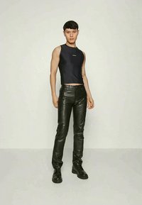 Jaunas vyras stovi juodame be rankovių marškinėlyje, juodose odinėse kelnių ir juoduose batuose ant paprasto šviesaus fono.