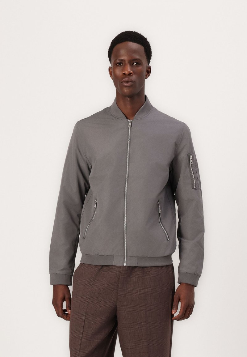 Veste bomber grise avec fermeture éclair à l'avant, deux poches latérales et une poche zippée sur la manche. Tissu lisse, poignets et ourlet côtelés.
