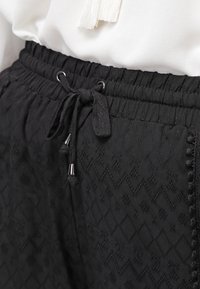 Pantaloni neri con coulisse in vita elastica, caratterizzati da un tessuto testurizzato con pattern geometrici. Finiti con dettagli in metallo argentato.