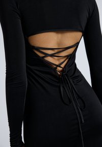 Robe noire à manches longues avec un dos ouvert, lacée de sangles croisées et nouée à la taille avec un ruban assorti.