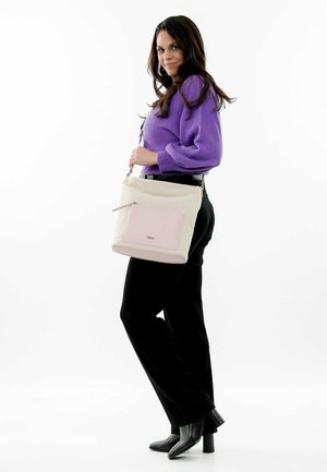 Sac seau léger beige et rose avec une poche zippée extérieure et un détail de logo, associé à un pull violet et un pantalon noir, sur fond blanc.