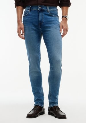 Mand iført slim fit blå jeans, sorte pæne sko, mørk skjorte og armbåndsur, stående på hvid baggrund.