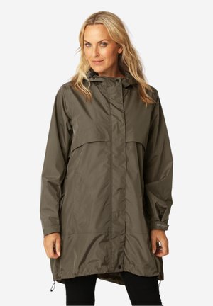 Blonde vrouw die een lange olijfgroene regenjas met kap is draagt, staand tegen een effen witte achtergrond.