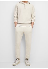 Beige hooded sweatshirt met een kangoeroezak, gecombineerd met bijpassende tapered joggingbroeken. Witte sneakers maken de outfit af. Glad stoffen textuur.