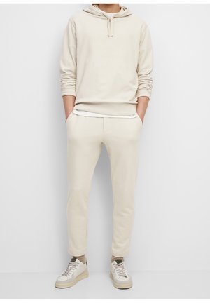 Beige Kapuzenpullover mit Kängurutasche, kombiniert mit passenden, schmal zulaufenden Jogginghosen. Weiße Sneaker runden das Outfit ab. Glatte Stoffstruktur.