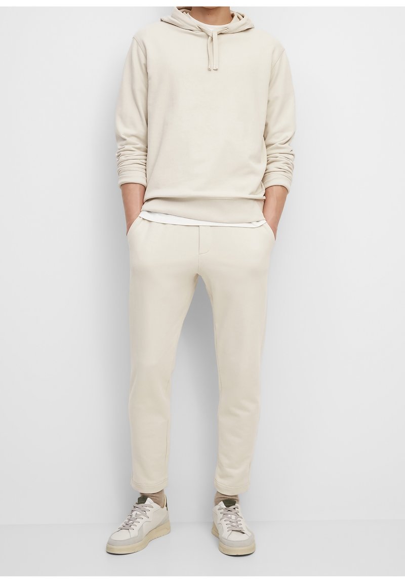 Beige hooded sweatshirt met een kangoeroezak, gecombineerd met bijpassende tapered joggingbroeken. Witte sneakers maken de outfit af. Glad stoffen textuur.
