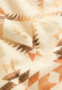 Textile présentant un fond crème avec des motifs géométriques en marron, orange et beige, composée de triangles et de flèches, mettant en valeur une texture douce.