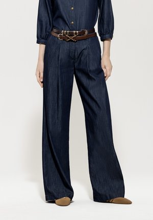 Weite, dunkle Jeans mit hoher Taille, ergänzt durch einen braunen Gürtel mit goldfarbener Hardware. Der Stoff hat eine strukturierte Oberfläche.
