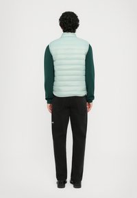 Uomo con capelli ricci che indossa un gilet trapuntato azzurro chiaro sopra una maglietta a maniche lunghe verde scuro, jeans neri e scarpe nere, in piedi con la schiena rivolta.