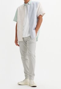 Man wearing pastel striped short-sleeve button-up shirt, white t-shirt, light gray pants, and white sneakers, standing with one hand in pocket.