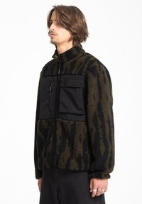 Fleece- ja nailonista valmistettu vetoketjullinen takki, jossa on oliivinvihreä ja musta eläinkuos, rintatasku, korkea kaulus ja teksturoitu kangas.