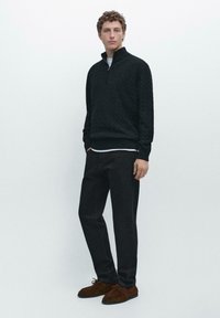 Maglione nero lavorato a maglia con colletto alto, abbinato a jeans scuri e scarpe in camoscio marrone. Presenta un motivo strutturato e una camicia bianca a strati sotto.