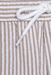 Tissu rayé avec des lignes blanches et marron clair alternées. Cordons ajustables blancs avec une pointe en métal. Matériau doux et texturé.