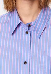 Light blue button-up shirt featuring vertical pink stripes, a pointed collar, and a small pocket on the left chest. Buttons are dark brown.