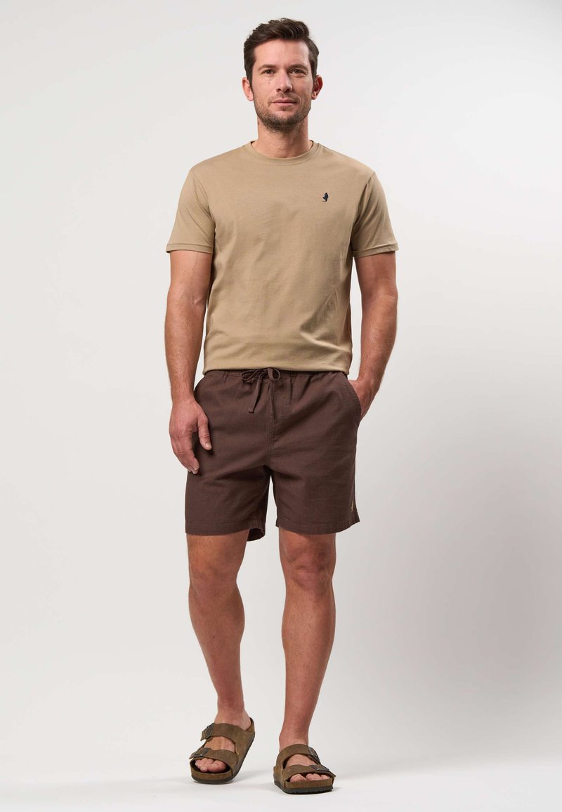 Homme portant une chemise beige à manches courtes, un short marron avec cordon de serrage et des sandales marron, debout devant un fond clair uni.