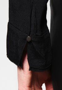 Manica in tessuto nero con texture, dotata di polsino con bottone e dettaglio a contrasto sull'orlo. Presenta cuciture visibili e leggero sfilacciamento ai bordi.