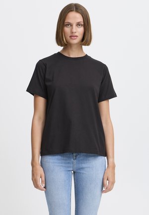 Žena s rovnými vlasmi po plecia, oblečená v jednoduchom čiernom tričku s krátkym rukávom a svetlomodrých džínsoch, stojaca pred bielym pozadím.