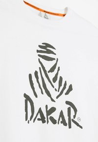Biała bawełniana koszulka z dużym, abstrakcyjnym, ciemnozielonym logo przypominającym zamaskowaną postać, z napisem "DAKAR" na dole.